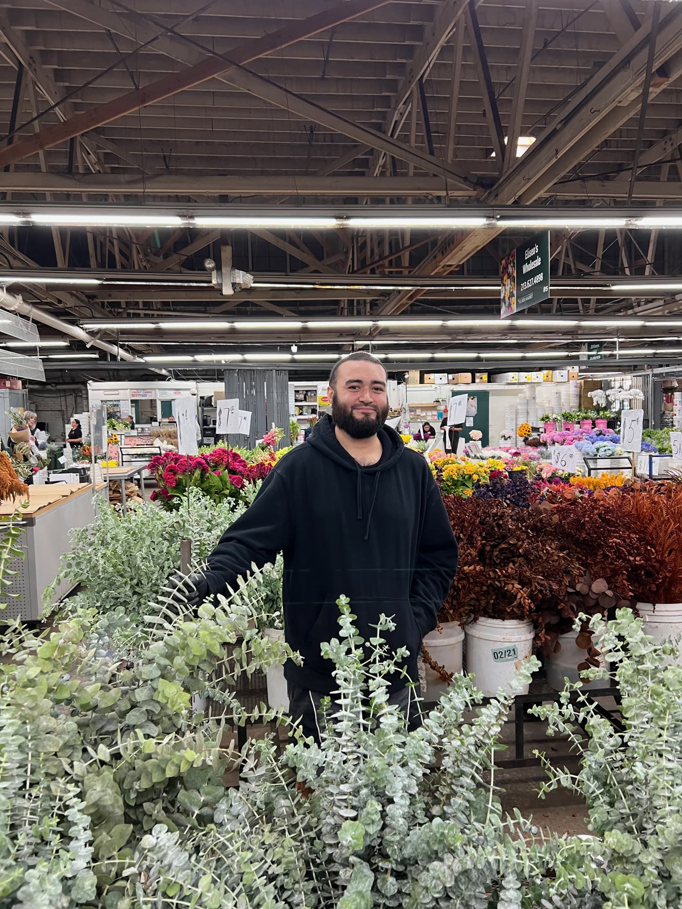 Anthony standing behind buckets of eucalyptus at stall 15 in the LA Flower Market, the Eliseo's Wholesale sign overhead